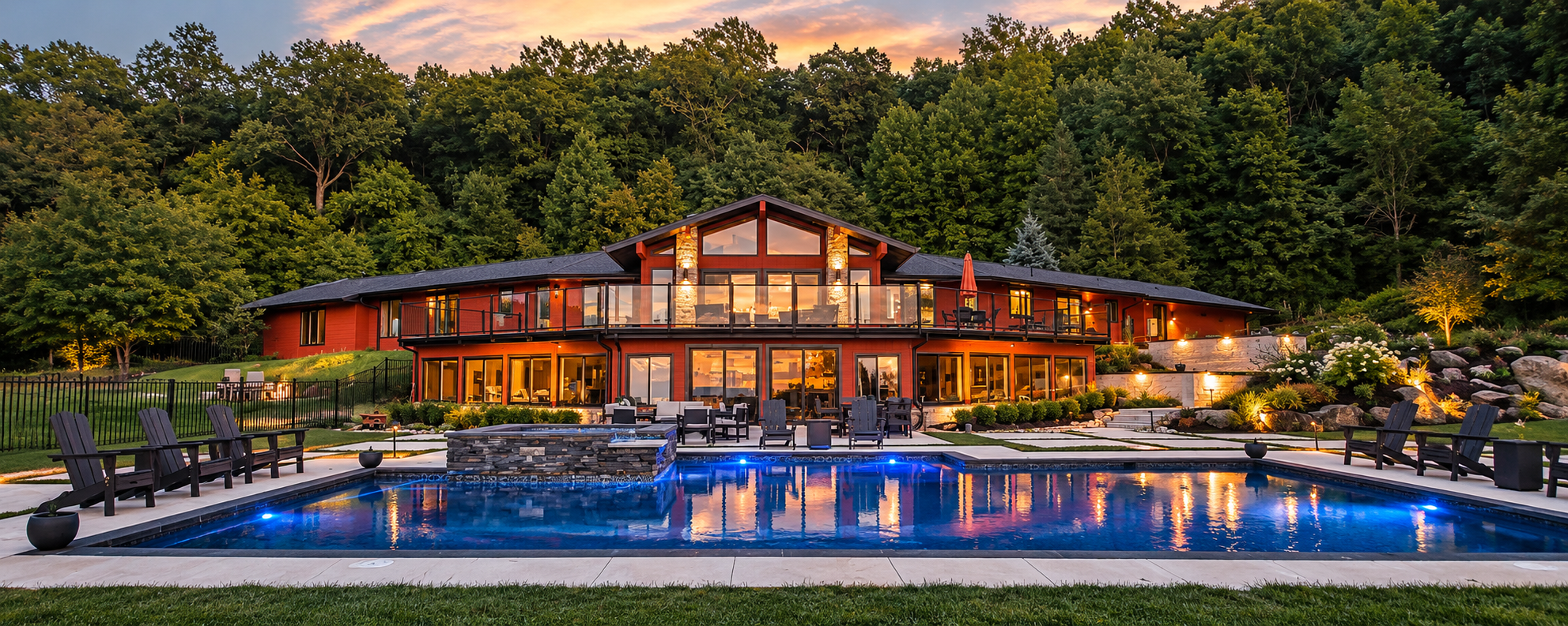 Renovated rear exterior of our 1980s home at dusk with an illuminated pool, expansive glass windows, outdoor living areas and wooded surroundings.
